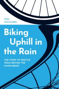 Book cover for "Biking Uphill in the Rain" by Tom Fulcoloro, featuring a minimalist illustration of a bicycle wheel and its distorted reflection in a large blue puddle.