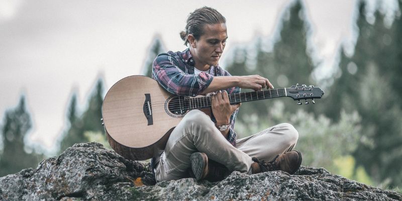 Calum Graham sits on a rock outdoors and tunes his acoustic guitar.