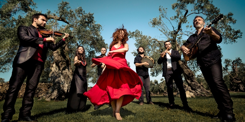 Canzoniere Greciano Salentino performs outdoors under a clear sky. 6 of the members are dressed in black and play instruments, while one member wears a red dress and dances in the center.