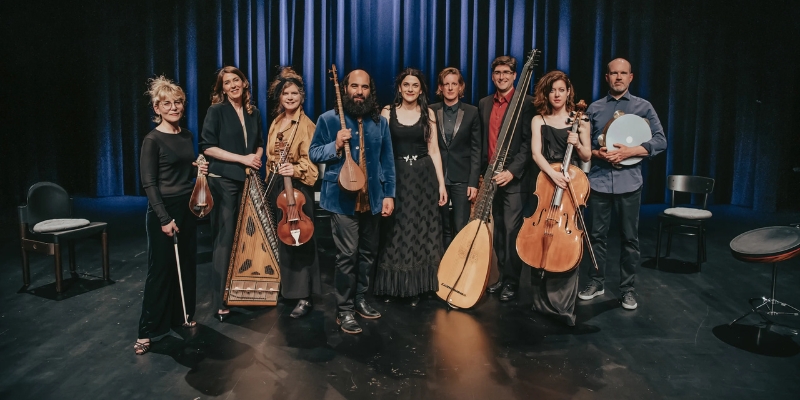 A co-ed string ensemble of 9 performers pose with their instruments in front of navy stage curtains.
