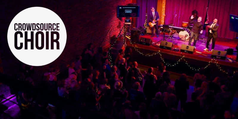 A small, intimate music venue with a live band on a stage, lit by stage lights and fairy lights. The audience sings along to lyrics shown on a TV screen while the conductor makes arm gestures. The Crowdsource Choir logo is in the upper left corner.