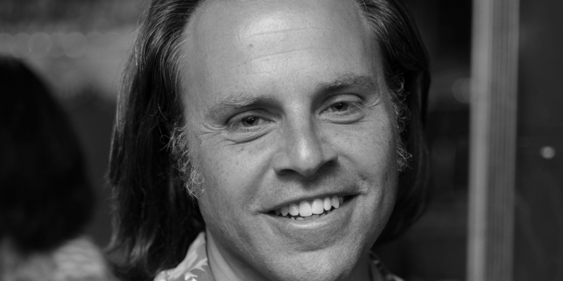 Black and white headshot of Daniel Brook (with light skin and chin-length hair) wearing an abstract print shirt.