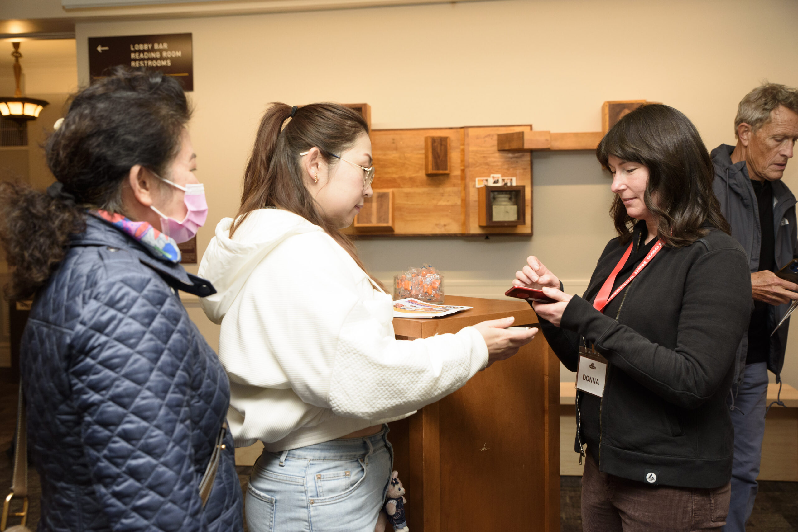 A volunteer in a black sweater with dark hair checks in two patrons one in a white sweater with glasses and the other in a dark jacket. 