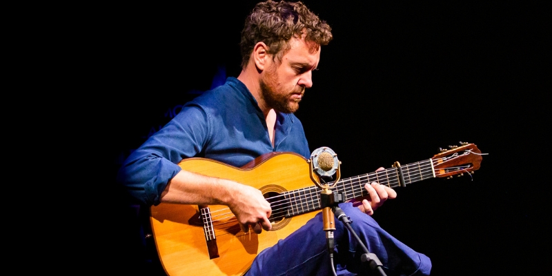 Derek Gripper wears a blue shirt and blue pants while playing an acoustic guitar on stage behind a microphone.