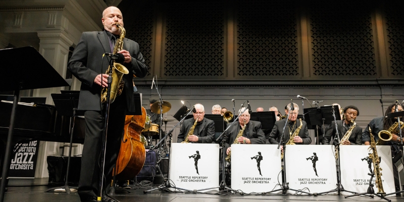 A saxophone player wearing an all black suit performs on stage with an accompanying band and orchestra behind him. "Seattle Repertory Jazz Orchestra" signs are placed in front of the orchestra.