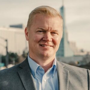 Headshot of Eli Arnold (with fair skin, short blonde hair, and grey suit)