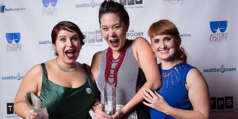 A trio dressed in formal clothing smiles and poses, with the person in the center holding a Gregory Award.