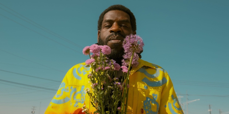 Hanif Abdurraqib (with dark skin, textured black hair & beard) poses with a bouquet of pink flowers in his hands.