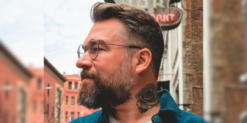 Headshot of Issac Fitzgerald (with short brown hair, beard, eyeglasses, and a rose neck tattoo)