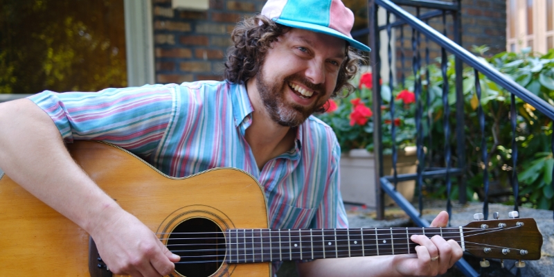 Jon Pontrello (with fair skin, wavy brown hair/beard) plays the guitar and smiles while wearing a colorful baseball cap and striped shirt.