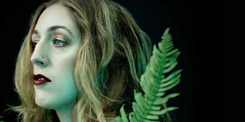 Headshot of Kaley Lane Eaton (with fair skin, wavy blonde hair, and dark lipstick) posing next to a fern leaf.