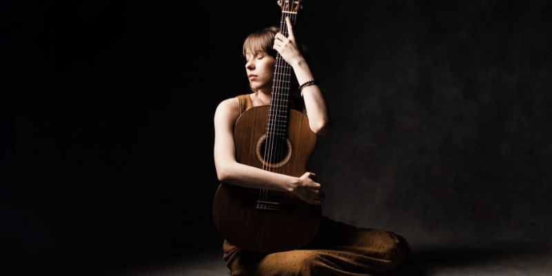 Kasia Smolarek (with fair skin and brown hair) sits in front of a black background and poses with her arms wrapped around an acoustic guitar.