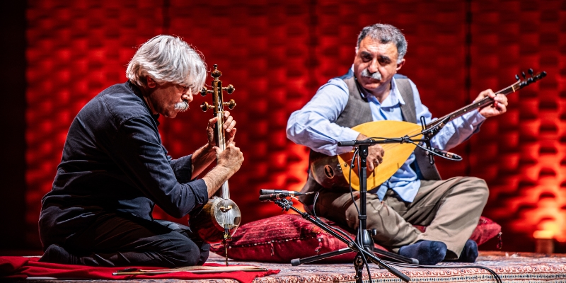 Kayhan Kalhor (playing the kamancheh) and Erdal Erzincan (playing the bağlama) perform together while sitting on a Persian rug. The stage is brightly lit, with red lights in the background.