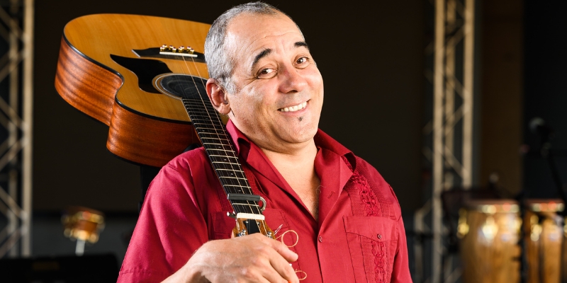 Kiki Valera (wearing a red button-down shirt) smiles and carries his acoustic guitar over one shoulder.