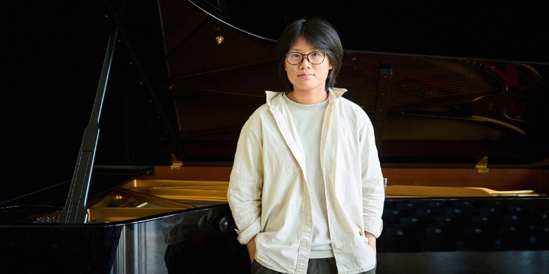Magdalene Ho (with short black hair, eyeglasses, and white button-down shirt) stands in front of a grand piano with her hands in her pockets.