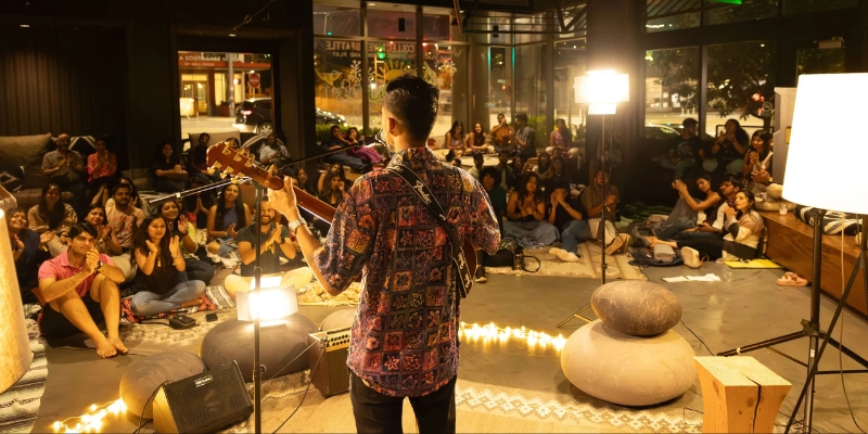 A person with short black hair and a patterned shirt plays the guitar on a small, warmly lit stage. The audience is seated on the floor, atop rugs and cushions.