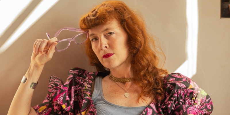 Headshot of Melissa Auf der Maur (with fair skin, wavy ginger hair, and red lipstick. She wears a colorful blouse with a gray tank top and gold necklaces.