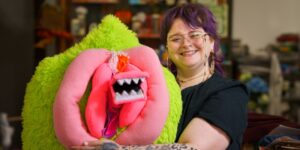 Mikki Ulaszewski (with purple hair and eyeglasses) smiles and holds up one of their puppets.