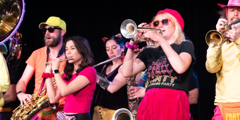 A small group of performers play various brass instruments on stage (flute, trumpets, tuba, saxophone) while wearing colorful neon clothing.