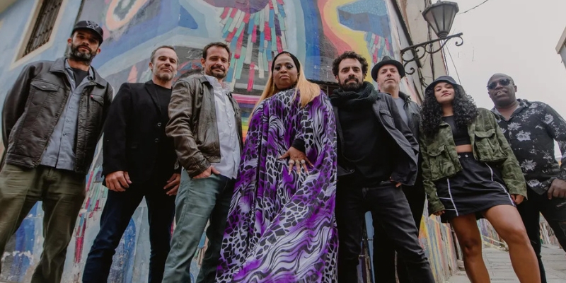 All 8 members of Novalima pose together in front of a colorful mural, looking down at the camera.