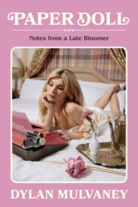 Book cover of "Paper Doll: Notes from a Late Bloomer" by Dylan Mulvaney, featuring a Dylan (with blonde hair and fair skin) reclining on a bed, gazing at a pink typewriter with a crumpled paper, a flower vase, and a silver tea set nearby. The background includes patterned curtains and a soft pink border with the title and author's name in a vintage font.