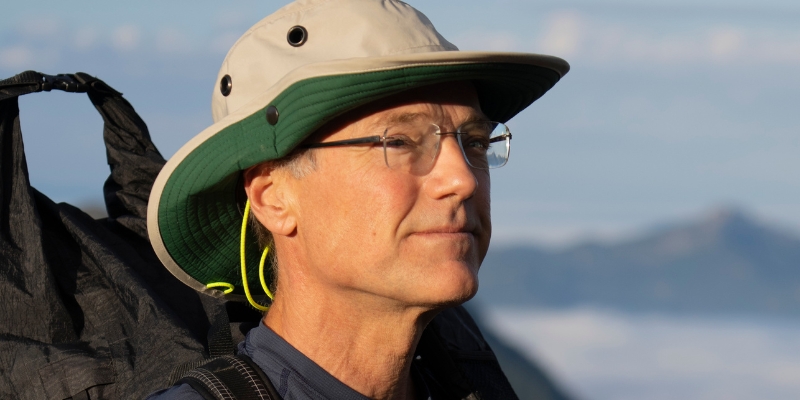 Outdoor headshot of Paul Bannick (with fair skin and eyeglasses) wearing a beige bucket hat and black backpack.