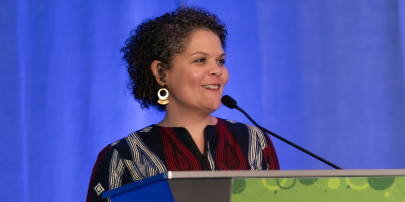 A person with curly black hair and a patterned blouse smiles and speaks in front of a podium.