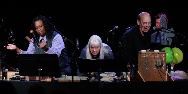Three performers stand behind a table and play instruments into microphones while reading sheet music.