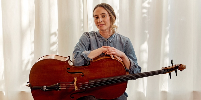 Ollella sits and poses in front of white sunlit curtains. Her arms are folded on top of her cello, which sits sideways in her lap.