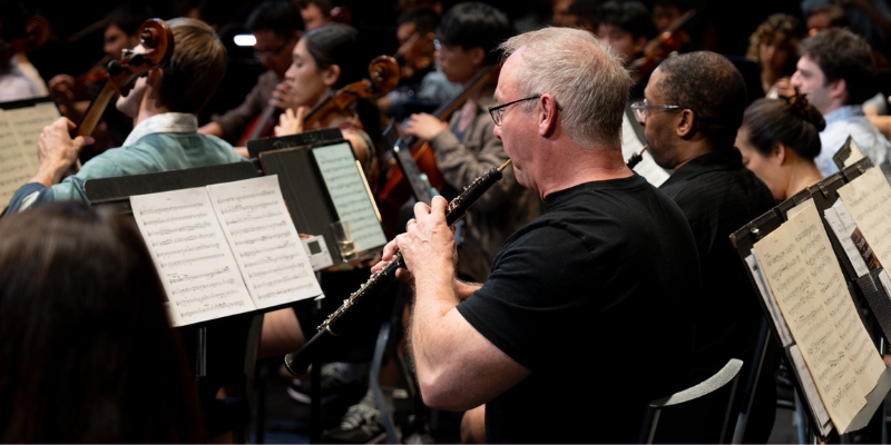 Close-up shot of a person wearing a black t-shirt and playing the clarinet in an orchestra.