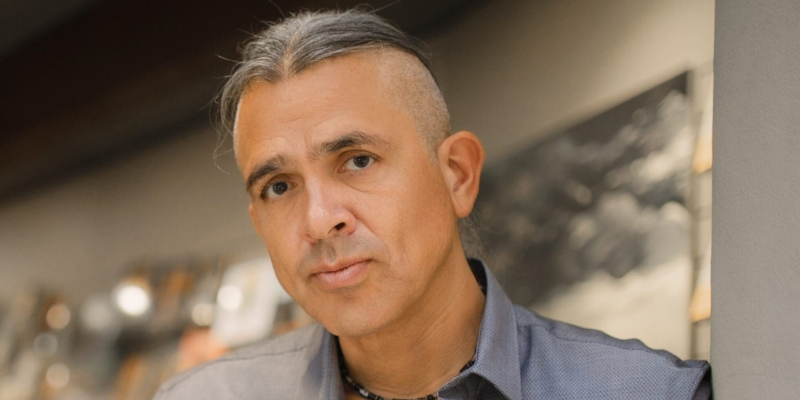 Headshot of Steven Graham Jones (with tan skin and grey hair tied back). The sides of his head are shaved.