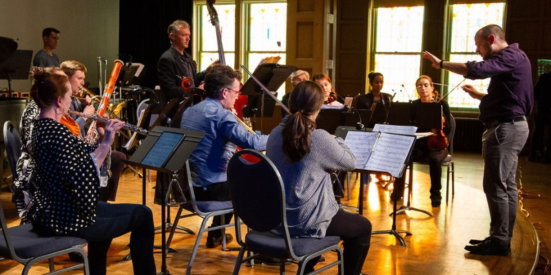 A small ensemble of musicians practice with a conductor. The room is naturally lit by sunlight, coming in from large windows.
