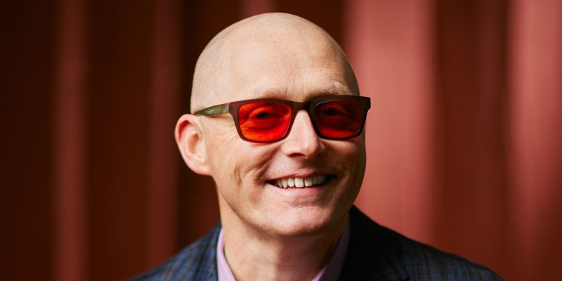 Headshot of Thomas Marriott (with shaved head, red sunglasses, and dark plaid suit)