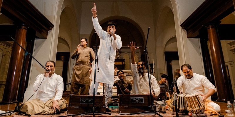 Six men perform in a room with arched walls and columns, playing traditional South Asian instruments and singing. In the center, one man stands wearing a white traditional outfit, singing into a microphone, and gesturing with one hand raised, pointing upward.
