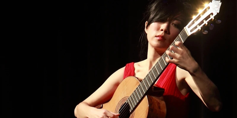 Xuefei Yang (with fair skin, red dress, and black bangs) plays the guitar in front of a black background.