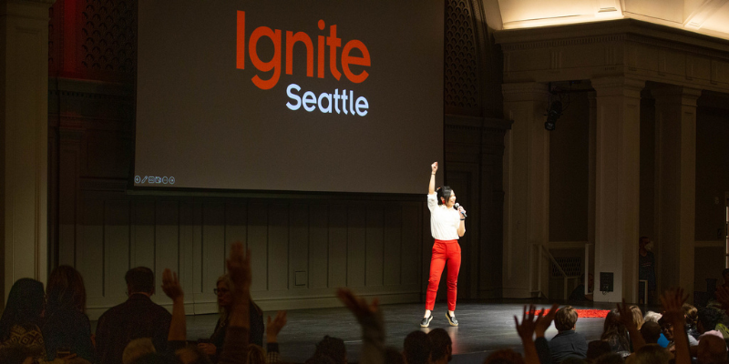 A person wearing a white shirt and red pants stands on stage with their fist raised, holding a microphone in the other hand. The Ignite Seattle logo is projected behind them.